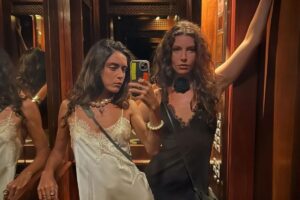 Alanis Guillen e Giovanna Reis posam juntas em frente ao espelho de um elevador com painéis de madeira. Alanis, à esquerda, usa um vestido branco de cetim com renda e um colar de pérolas grandes. Giovanna, à direita, segura o celular, veste um vestido preto com detalhes em renda e uma flor preta no pescoço.