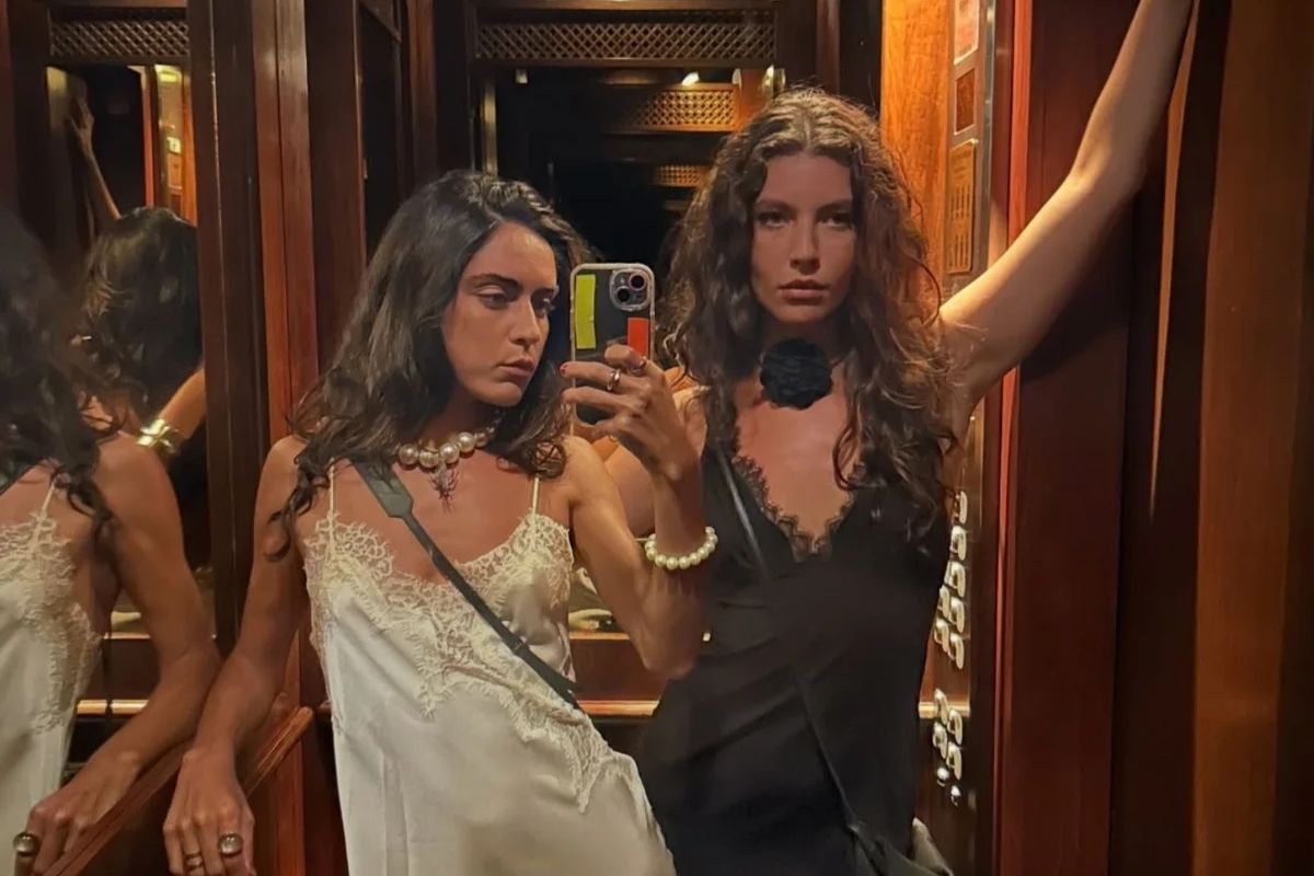 Alanis Guillen e Giovanna Reis posam juntas em frente ao espelho de um elevador com painéis de madeira. Alanis, à esquerda, usa um vestido branco de cetim com renda e um colar de pérolas grandes. Giovanna, à direita, segura o celular, veste um vestido preto com detalhes em renda e uma flor preta no pescoço.
