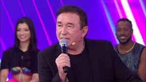 Cantor Amado Batista de terno preto segura microfone prateado enquanto se apresenta no palco do Programa Silvio Santos, no SBT. Ao fundo, dançarinos sorriem em um cenário com luzes vibrantes em tons de roxo e rosa.