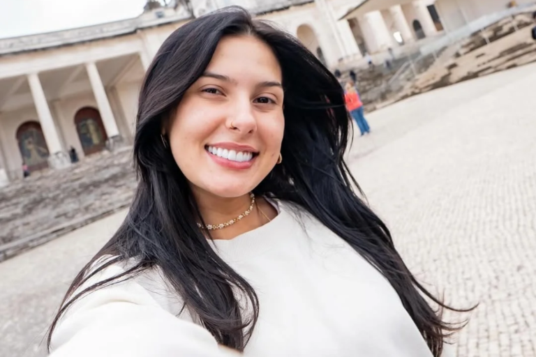Selfie da cantora Ana Castela sorrindo ao ar livre. Ela possui cabelos longos e pretos, usa uma blusa branca de manga comprida, um colar de flores delicado e um pequeno piercing no nariz. Ao fundo, uma construção histórica com colunas e escadarias sob luz natural.