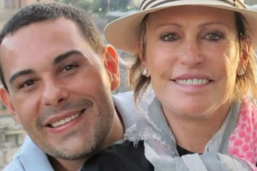 Close lateral de Ana Maria Braga sorrindo ao lado de Marcelo Frisoni. A apresentadora usa chapéu bege com faixa listrada, cachecol cinza e lenço rosa, enquanto Marcelo sorri com barba curta e camisa polo azul-clara. O fundo está desfocado.