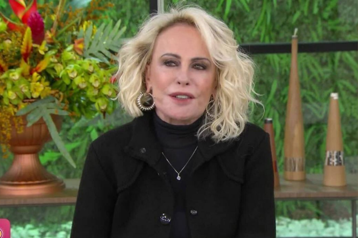 Ana Maria Braga sorrindo, com cabelos loiros cacheados e vestindo um casaco preto, em um estúdio de televisão com fundo verde e plantas.