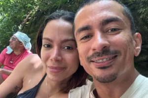 Selfie em close-up da cantora Anitta sorrindo suavemente e seu irmão Renan Machado rindo, ambos próximos um do outro em um ambiente natural com folhagens verdes ao fundo. Ao longe, um terceiro homem com turbante azul e camisa rosa observa de perfil.
