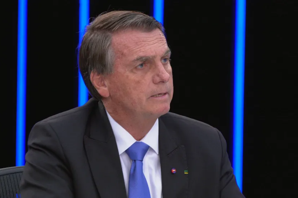 Fotografia em close de Jair Bolsonaro, um homem de pele clara e cabelos grisalhos curtos, usando um terno preto, camisa branca e gravata azul vibrante. Ele está com a cabeça levemente inclinada para a direita, com uma expressão séria e olhar atento. O fundo é composto por painéis pretos intercalados com linhas de luz azul vertical.