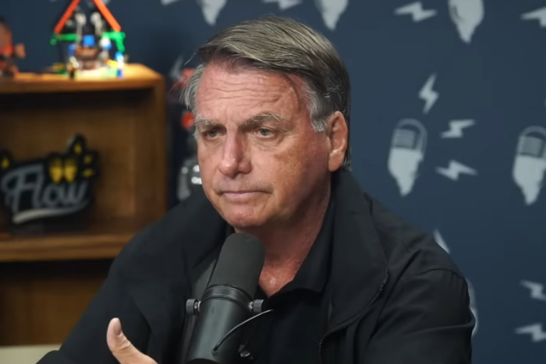 Jair Bolsonaro, ex-presidente do Brasil, de perfil lateral em entrevista ao Flow Podcast. Ele usa uma jaqueta preta e gesticula com a mão esquerda à frente de um microfone profissional. O fundo azul exibe a logomarca do programa com ilustrações de microfones e raios.