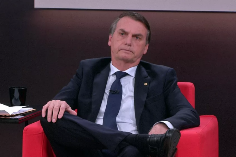 Jair Bolsonaro sentado em uma poltrona vermelha durante uma entrevista. Ele veste terno azul escuro, camisa branca e gravata azul. Está com as pernas cruzadas e mantém uma expressão facial séria. Ao fundo, um cenário de estúdio com tons escuros e uma mesa lateral com papéis e um copo preto.
