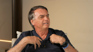 Jair Bolsonaro, ex-presidente do Brasil, sentado em uma poltrona clara. Ele veste uma camisa polo azul marinho, usa um relógio digital robusto no pulso esquerdo e gesticula com as mãos próximas ao peito enquanto fala. O fundo é neutro em tons de bege.