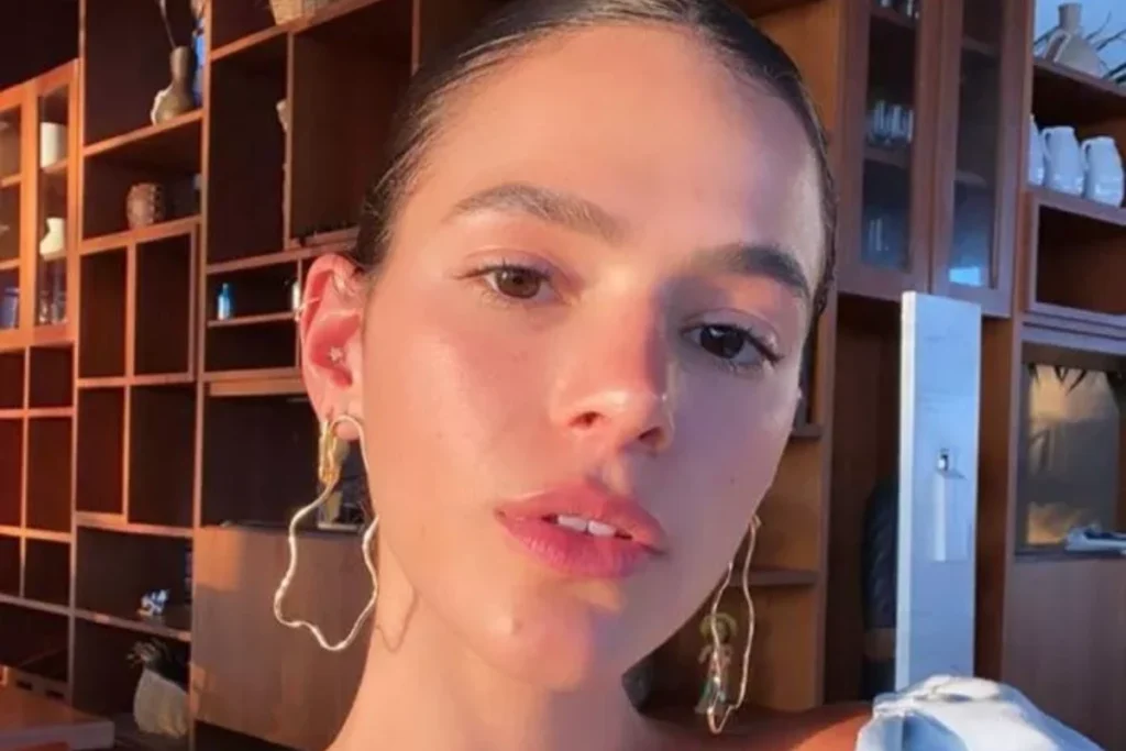 A atriz Bruna Marquezine aparece em um close-up frontal, olhando para a câmera com expressão séria e lábios levemente abertos. Ela está com o cabelo preso para trás, maquiagem natural e usa um brinco de argola dourada grande e escultural, em formato ondulado abstrato. O fundo é um ambiente interno com estantes de madeira repletas de objetos e uma janela ao longe, sob uma luz quente e dourada do pôr do sol.