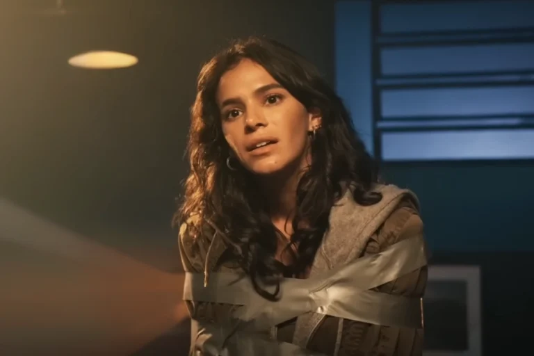 Atriz Bruna Marquezine, com expressão séria e surpresa, está sentada em um ambiente escuro, com fitas adesivas de metal enroladas ao redor de seu corpo e braços, sobre um casaco cinza com capuz. Seu cabelo está ondulado e há um ponto de luz ao fundo.