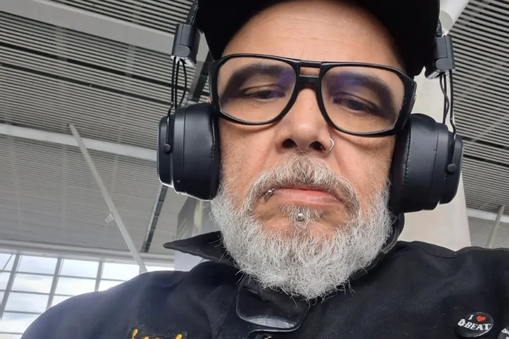 João Gordo em close, usando boné, óculos de grau grossos, fones de ouvido grandes e com a barba grisalha em destaque, posando com expressão séria. É possível ver seus piercings no nariz e no lábio.