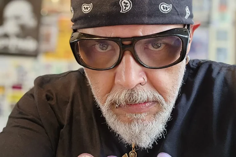 Close em João Gordo olhando diretamente para a câmera, usando bandana preta com estampa branca, óculos escuros com armação grossa e exibindo sua barba grisalha e piercings no nariz e lábio.