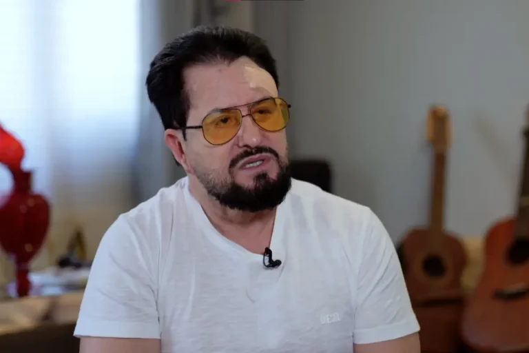 Marrone, da dupla Bruno e Marrone, com óculos de sol amarelos, camiseta branca e barba, olhando para a esquerda durante uma entrevista em um ambiente interno.