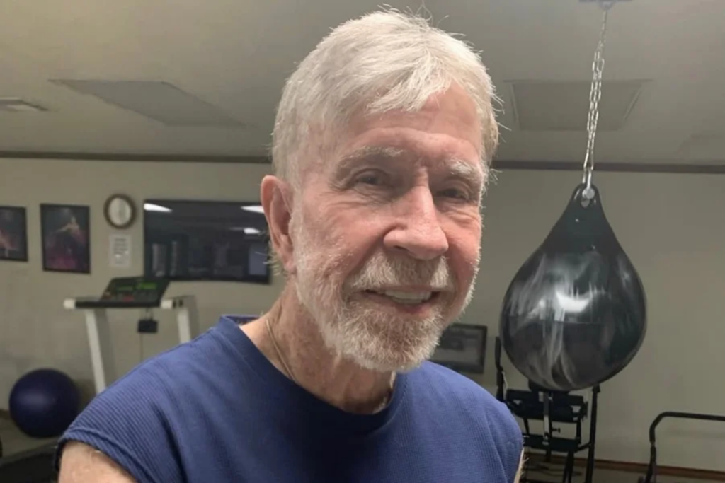 Ator Chuck Norris, um homem idoso, de cabelo e barba brancos e grisalhos, sorri para a câmera vestindo uma camiseta azul de malha canelada. Ele está em um ambiente de ginásio com equipamentos de exercício ao fundo.