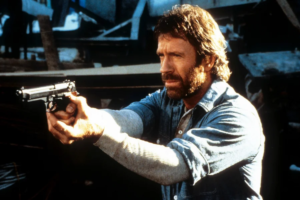 Homem de cabelo castanho e barba, vestindo camisa jeans sobre uma manga térmica branca, segura uma pistola preta com as duas mãos em pose de mira. O fundo é escuro e desfocado com estruturas industriais.