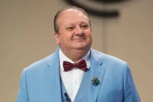Retrato em plano médio do chef de cozinha Erick Jacquin sorrindo. Ele veste um terno completo de cor azul claro, colete combinando, camisa social branca e uma gravata borboleta texturizada de cor bordô. No paletó, há um broche cravejado de pedras turquesas. Ao fundo, uma estrutura de madeira clara levemente desfocada.