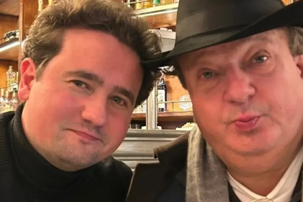 Erick Jacquin, chef de cozinha e jurado do MasterChef Brasil, de chapéu preto e cachecol cinza, ao lado de seu filho mais velho, Edoard Jacquin, que veste uma blusa de gola alta preta. Ambos estão em um ambiente interno que lembra um restaurante ou bar com prateleiras de garrafas ao fundo.
