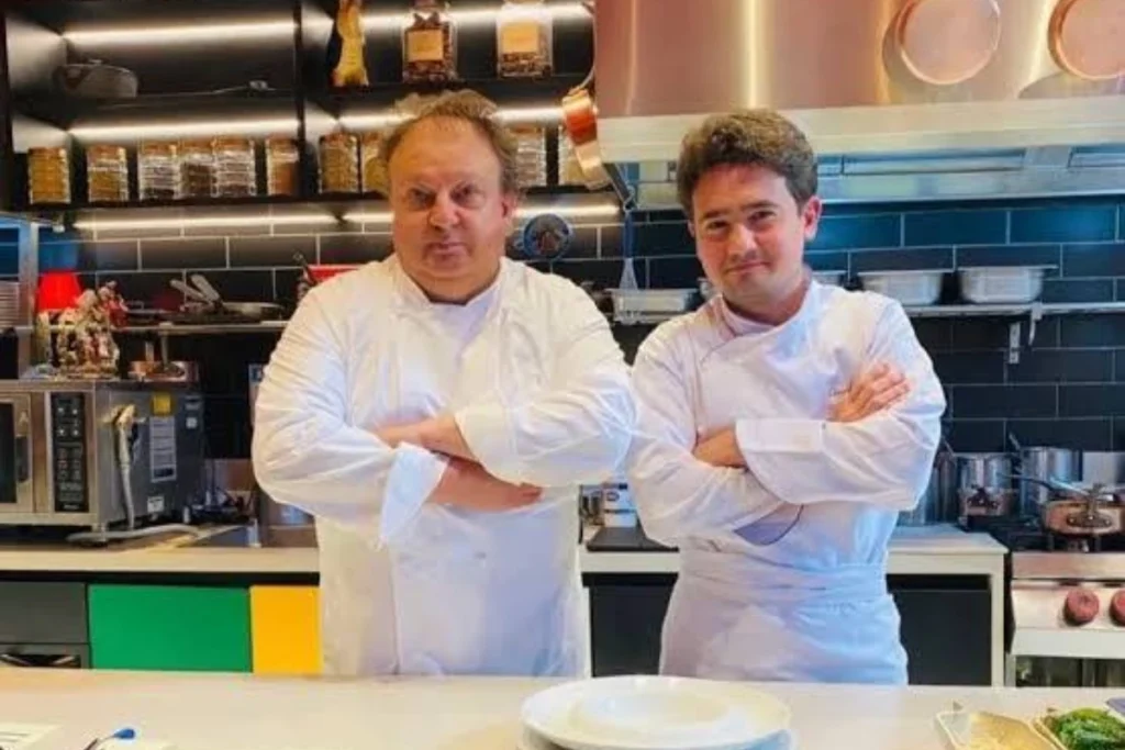Erick Jacquin e seu filho Edoard Jacquin posam de braços cruzados dentro de uma cozinha profissional. Ambos vestem dólmãs brancos de chef. Ao fundo, prateleiras com mantimentos, azulejos pretos e equipamentos de cozinha industrial.