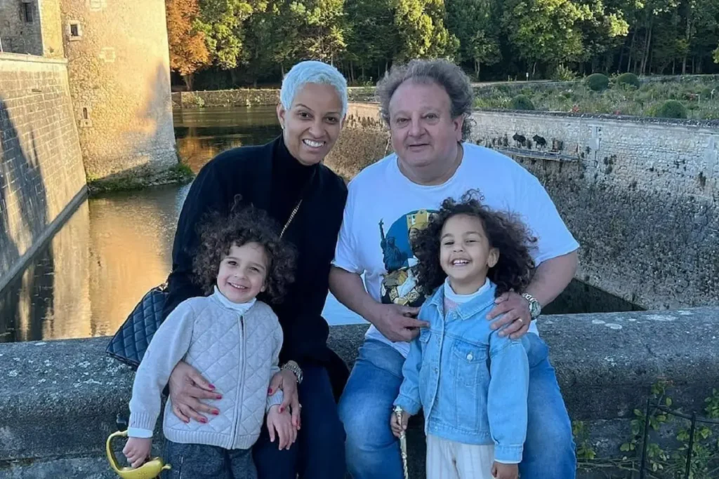 Erick Jacquin, chef de cozinha e jurado do MasterChef, de camiseta branca e calça jeans, sentado ao lado de sua esposa Rosângela Menezes, que tem cabelo curto e prateado e veste preto. À frente deles estão seus filhos gêmeos, Elise e Antoine, ambos com cabelos cacheados. Eles estão posando em uma ponte de pedra sobre um canal de água, com uma estrutura histórica de pedra e árvores ao fundo durante o entardecer.