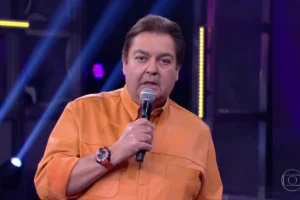 O apresentador Faustão segura um microfone próximo à boca enquanto fala. Ele veste uma camisa de botão laranja vibrante e usa um relógio de pulso grande com pulseira vermelha. O fundo mostra luzes de estúdio em tons de azul e roxo.