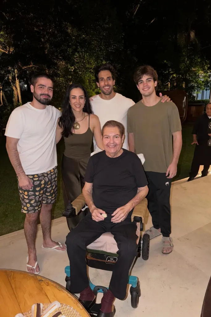 Faustão sentado em uma cadeira de rodas, sorridente, rodeado por seus três filhos — Lara, João Guilherme e Rodrigo — e o genro, Julinho Casares. Eles estão em uma área externa à noite, com árvores ao fundo e iluminação suave.