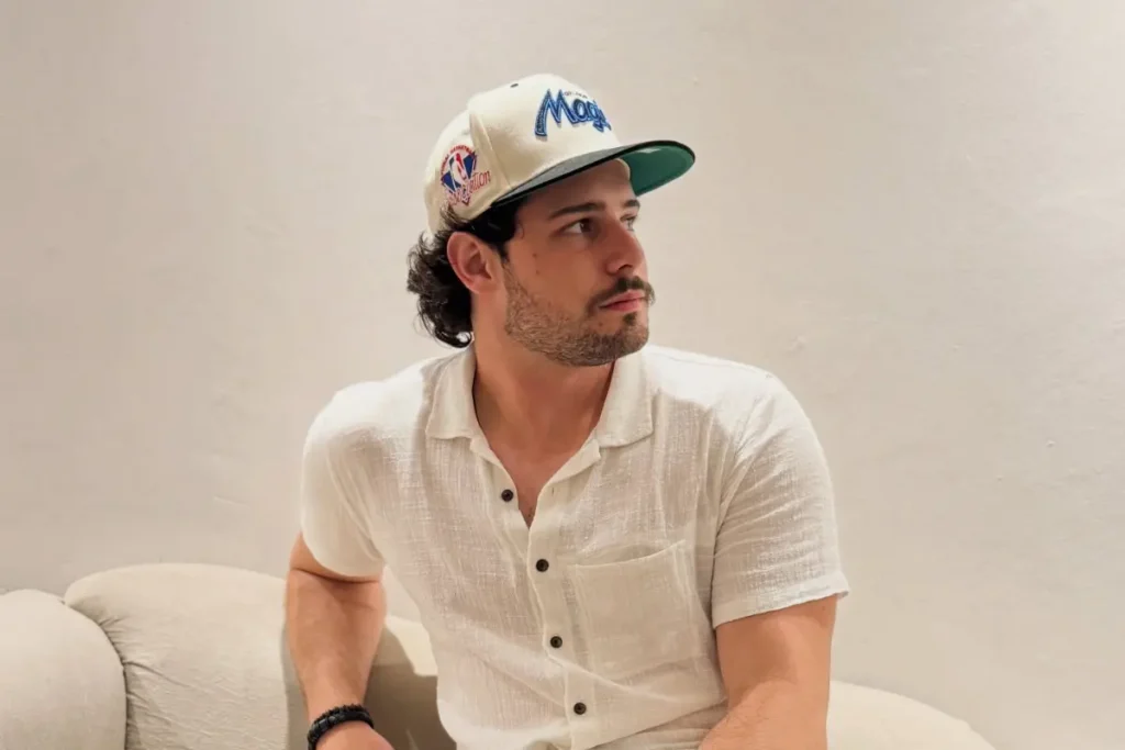 Homem jovem de pele clara, com barba rala e cabelos escuros cacheados, usando um boné branco com detalhes em azul e verde. Ele veste uma camisa de botão branca de manga curta e está sentado em um sofá claro, olhando para o lado.