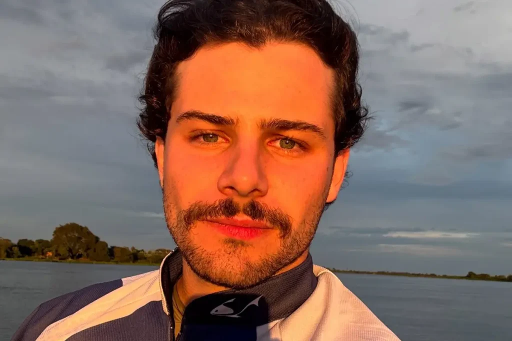 Homem jovem de pele clara, olhos claros e barba com bigode marcante, iluminado pela luz alaranjada do pôr do sol. Ele veste uma camisa esportiva azul e branca, com um rio e vegetação ao fundo sob um céu nublado.