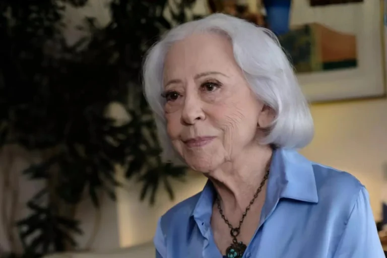 Fernanda Montenegro, uma mulher idosa com cabelo branco e curto, sorri levemente em um close-up, vestindo uma camisa de botão azul claro e um colar ornamentado.
