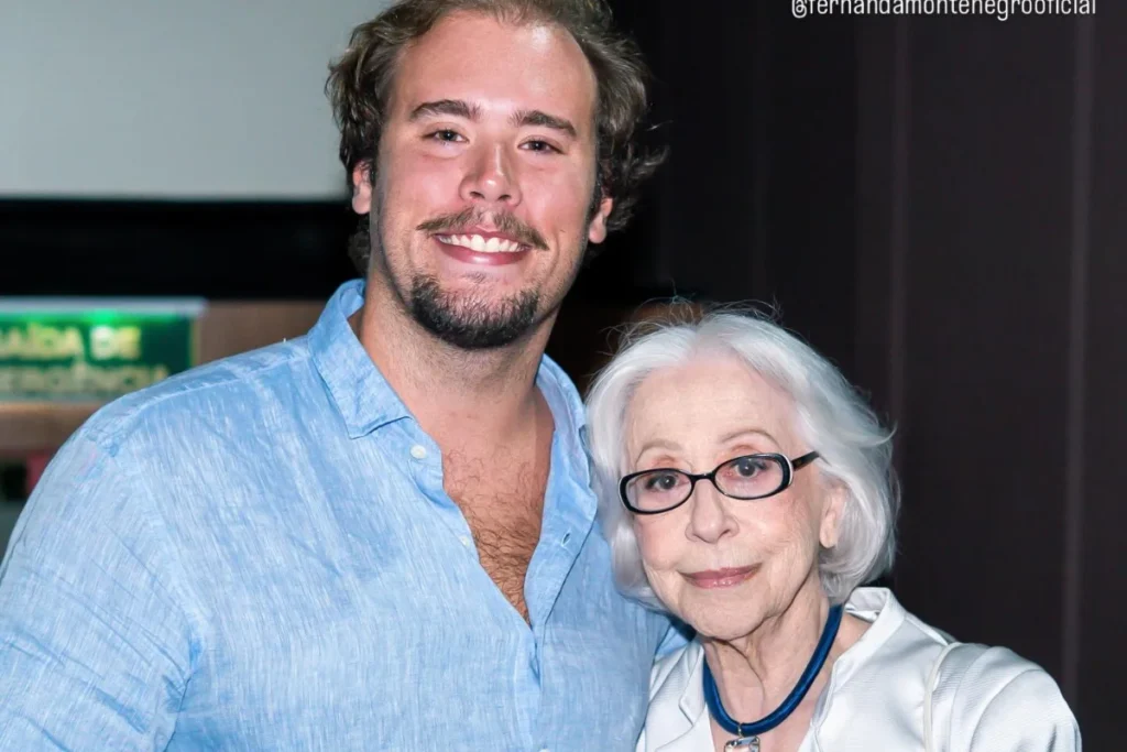 Fernanda Montenegro, usando óculos de grau e blusa branca, sorri ao lado de seu neto Davi Torres, que veste uma camisa de linho azul clara, em um registro afetuoso.