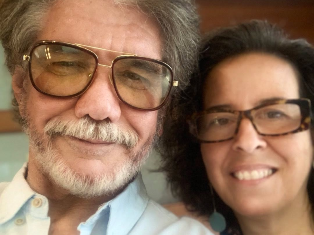 O ator Jackson Antunes, com cabelo grisalho e óculos escuros de lentes amareladas, sorri em uma foto selfie com sua esposa Cristiana Britto, que usa óculos de grau e brinco turquesa.