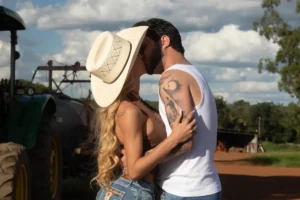 Gabi Martins, usando chapéu de cowboy bege e blusa marrom, beijando o influenciador Fidelis Falante em uma fazenda. Ao fundo, um trator verde e vegetação sob o céu ensolarado.