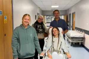 Holly Hubert sorri sentada em uma cadeira de rodas em um corredor de hospital, acompanhada por três familiares ou amigos que estão de pé ao seu redor. Ela usa uma bata hospitalar e possui tranças no cabelo.