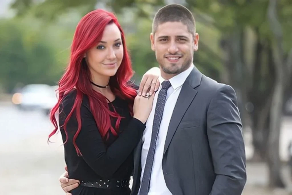 A atriz Josie Pessôa, com seu icônico cabelo longo vermelho vibrante e roupa preta, abraçada ao ator Daniel Rocha, que usa um terno cinza, camisa branca e gravata. Eles estão caracterizados como os personagens Du e João Lucas da novela Império, em um cenário externo diurno com árvores ao fundo.