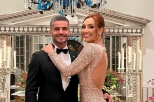 A atriz Josie Pessôa, ruiva e sorridente, usa um vestido de gala longo com paetês dourados e decote nas costas. Ela abraça seu marido, Luiz Danielian, que veste um smoking preto clássico com gravata borboleta. Eles estão em um ambiente sofisticado com um lustre azul e castiçais acesos ao fundo.