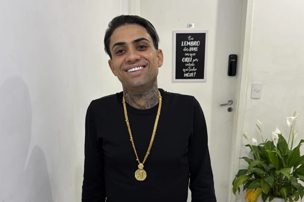Jota Surfista sorrindo de pé em um corredor. Ele tem cabelo escuro curto, está usando uma blusa de manga longa preta e uma corrente de ouro grossa com um pingente de cifrão. Tatuagens são visíveis em seu pescoço. O fundo mostra uma porta com um quadro preto com letras brancas e uma planta de copo-de-leite.