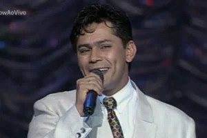 O cantor sertanejo Leandro, da dupla Leandro & Leonardo, canta sorrindo em um palco, segurando um microfone próximo à boca. Ele veste um paletó branco texturizado, camisa social branca e uma gravata colorida com padrão geométrico.