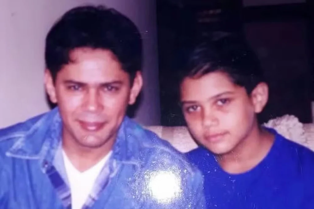 O cantor sertanejo Leandro, da dupla Leandro & Leonardo, sorri ao lado de seu filho Thiago Costa ainda criança. Leandro veste uma jaqueta jeans sobre camiseta branca e Thiago usa uma camiseta azul. A foto possui textura de fotografia analógica antiga.