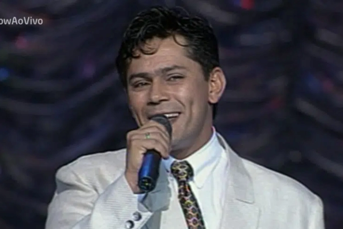 O cantor sertanejo Leandro, da dupla Leandro & Leonardo, canta sorrindo em um palco, segurando um microfone próximo à boca. Ele veste um paletó branco texturizado, camisa social branca e uma gravata colorida com padrão geométrico.