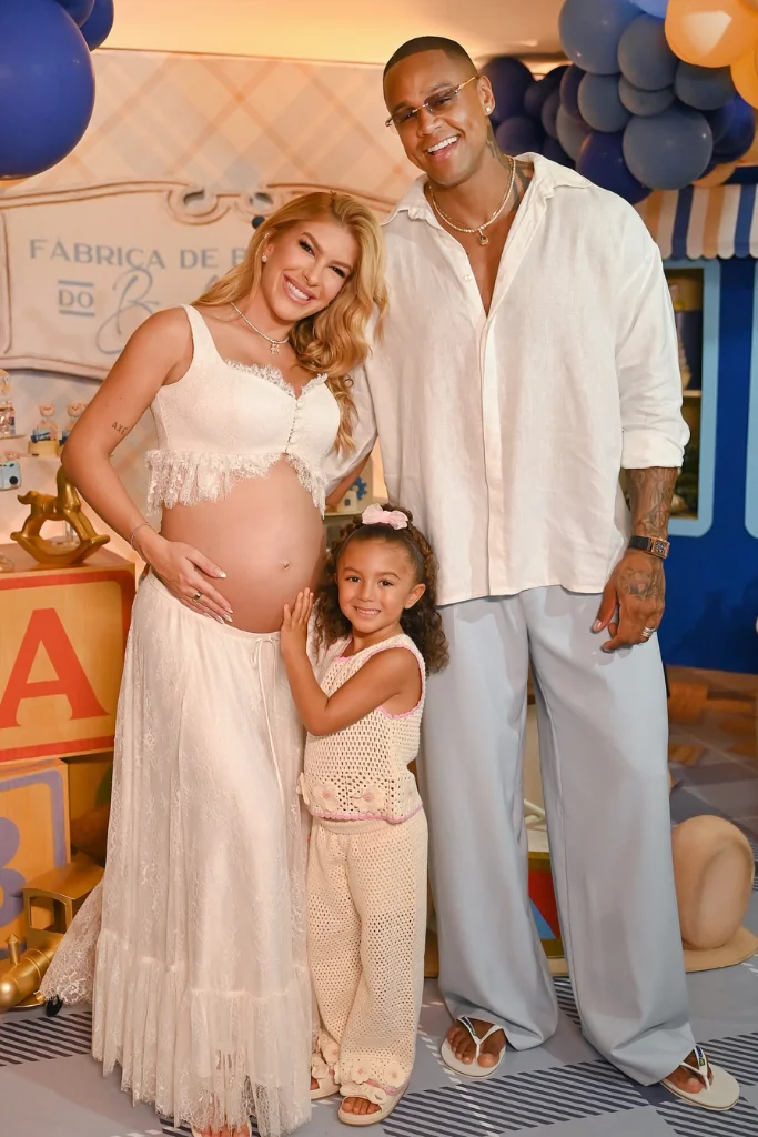 Lore Improta, grávida e usando um conjunto de renda branco que deixa a barriga à mostra, sorri ao lado de Léo Santana, que veste camisa branca e calça clara. À frente deles, a filha pequena do casal, Liz, sorri enquanto toca suavemente a barriga da mãe em um cenário de festa infantil com balões azuis.