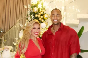 Lore Improta, mulher loira de cabelos longos, e Léo Santana, homem negro de cabelos curtos, ambos sorrindo e vestindo roupas vermelhas. Eles estão posicionados à frente de uma árvore de Natal decorada com bolas douradas e brancas, em um ambiente interno com escada ao fundo.