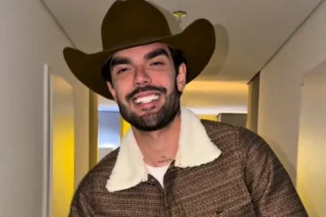 Luan Pereira sorridente usando um chapéu de cowboy marrom, jaqueta texturizada com gola de pelos branca e exibindo uma tatuagem no pescoço escrita "Gratidão".