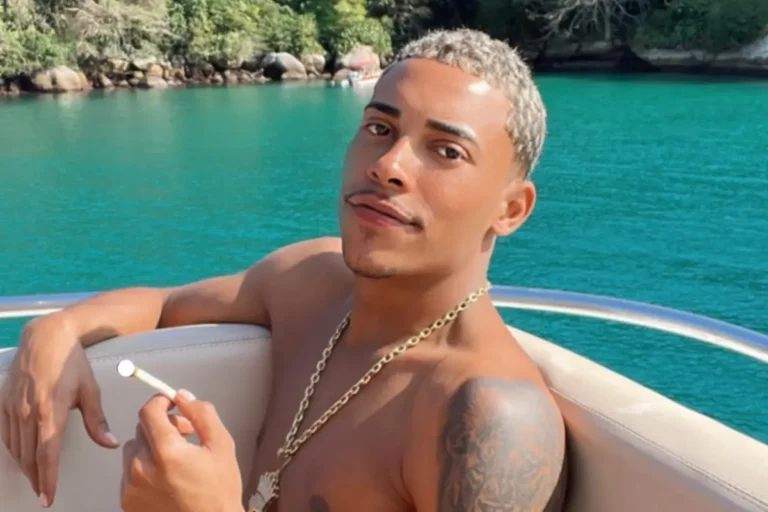 Homem jovem de pele retinta e cabelos curtos descoloridos de loiro, sem camisa, usando cordão de ouro grosso, sentado em um barco em alto-mar com águas esverdeadas e vegetação ao fundo. Ele segura um cigarro artesanal entre os dedos.