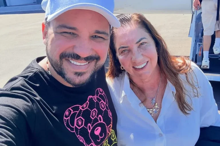 Margareth Serrão e Danilo Nascimento sorriem em uma selfie ao ar livre, em frente a uma aeronave. Margareth usa uma camisa branca e colares, enquanto Danilo veste uma camiseta preta com estampa de urso rosa e boné branco.