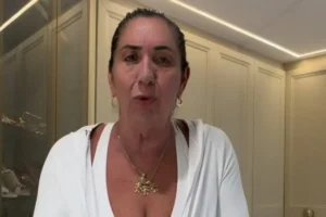 Margareth Serrão, mãe da influenciadora Virginia Fonseca, de frente para a câmera, usando uma blusa branca com decote em V, colares dourados e brincos de argola em um ambiente com armários claros ao fundo.