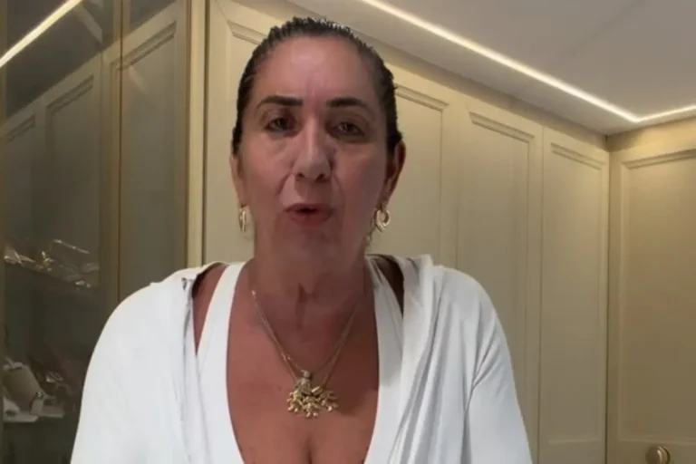 Margareth Serrão, mãe da influenciadora Virginia Fonseca, de frente para a câmera, usando uma blusa branca com decote em V, colares dourados e brincos de argola em um ambiente com armários claros ao fundo.