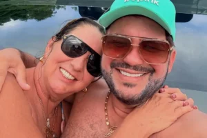 Margareth Serrão e Danilo Nascimento sorrindo abraçados em um ambiente ao ar livre. Margareth usa óculos de sol escuros e acessórios dourados, enquanto Danilo veste um boné verde claro, óculos de sol de lentes marrons e uma corrente dourada.