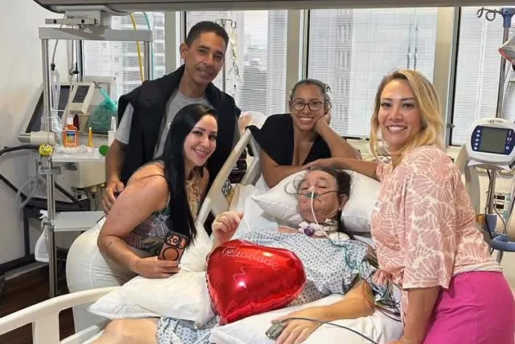 O humorista Marquito deitado em uma cama de hospital, usando avental médico e recebendo auxílio de oxigênio, cercado por quatro familiares sorridentes. Ele segura um balão vermelho em formato de coração com a palavra Felicidade escrita. O ambiente é um quarto de hospital moderno com equipamentos médicos ao fundo.