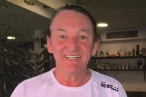 Retrato em close do humorista Marquito, sorrindo e olhando para a câmera, com o cabelo preso. Ele veste uma camiseta branca de gola redonda que tem um pequeno logotipo do "Grupo M&O" no peito. O fundo é um interior ligeiramente desfocado, mostrando prateleiras com garrafas e objetos variados, e algumas plantas verdes.