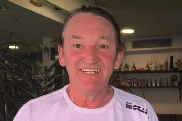 Retrato em close do humorista Marquito, sorrindo e olhando para a câmera, com o cabelo preso. Ele veste uma camiseta branca de gola redonda que tem um pequeno logotipo do "Grupo M&O" no peito. O fundo é um interior ligeiramente desfocado, mostrando prateleiras com garrafas e objetos variados, e algumas plantas verdes.