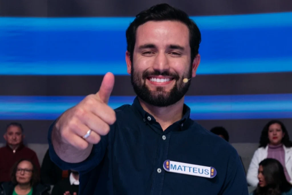 Matteus Amaral, ex-BBB, sorri e faz um gesto de 'joinha' com o polegar para a câmera em um estúdio de televisão, usando uma camisa azul escura e um crachá com o nome 'MATTEUS'.