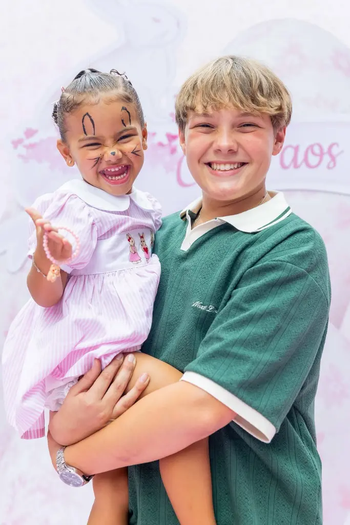 Davi Lucca, sorridente e vestindo uma camisa polo verde, segura no colo sua irmã caçula, Mavie. A bebê usa um vestido rosa e branco com detalhes de coelhinhos e está com o rosto pintado de coelho, com bigodes e orelhas desenhados em preto.